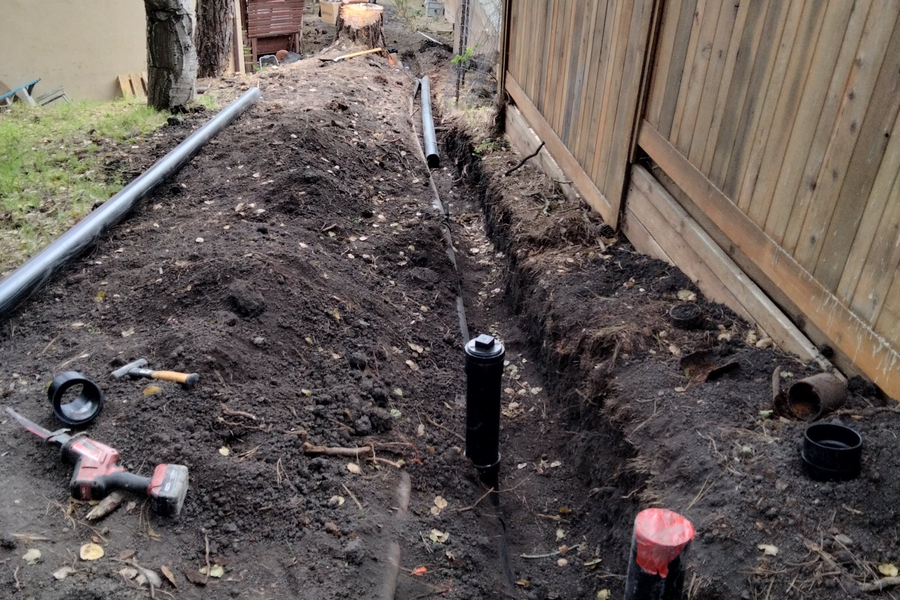 An open trench running along a side-yard fence with a new black sewer line laid in it and two cleanout risers set on the run, the old clay pipe pulled out beside the spoil